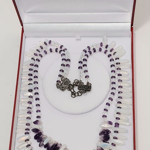 May include: A two-strand necklace with a silver clasp, displayed in a red box. The necklace features white and purple beads, with a mix of elongated white and dark purple stones. The necklace is designed with a layered effect.