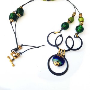 May include: A handcrafted necklace featuring a mix of green and black beads, gold accents, and a unique pendant. The pendant has a blue and gold design, suspended within black metal rings. The necklace has a toggle clasp.