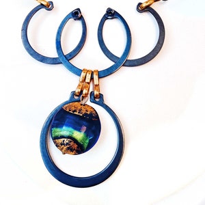 May include: A handcrafted necklace featuring a dark blue metal pendant with a circular design, accented by a dichroic glass bead with gold leaf. The necklace includes green glass beads, leather cord, and gold-toned metal accents. The clasp is a gold-toned toggle.