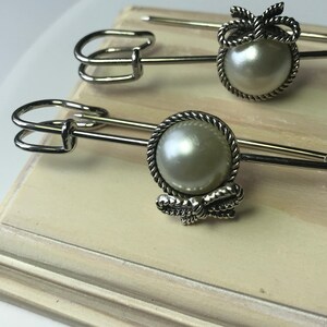 May include: Two silver-toned safety pins with pearl-like centers and decorative bow accents. The pins are displayed on a light-colored surface, showcasing their elegant design. The pins are likely used for fashion or craft projects.