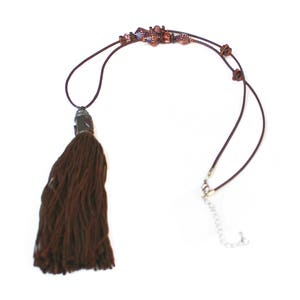 May include: A brown tassel necklace with a decorative beaded section and a clasp closure. The necklace features a long, dark brown tassel pendant and a series of small, square beads in shades of purple and brown. The necklace is on a white background.