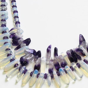 May include: A layered necklace featuring a heart-shaped design. The necklace combines white, teardrop-shaped beads with purple, rounded beads. The necklace is secured with a silver clasp. The necklace is a piece of jewelry.