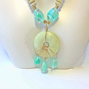 May include: A necklace featuring a large, light green circular pendant with a central hole, accented with gold wire and dangling teardrop-shaped beads. The necklace includes light blue and clear beads, and wooden beads.