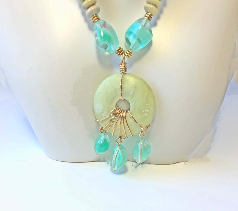 May include: A necklace featuring a large, light green circular pendant with a central hole, accented with gold wire and dangling teardrop-shaped beads. The necklace includes light blue and clear beads, and wooden beads.