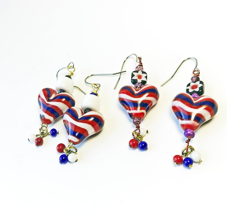 May include: Heart-shaped earrings with red, white, and blue stripes. The glass earrings have a dangling cluster of red, white, and blue beads. The earrings have a gold-colored wire and a white bead at the top.