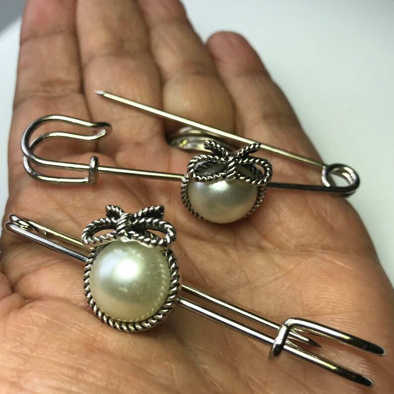May include: Three silver-toned safety pins with decorative pearl accents. Each pin features a large, round, off-white pearl set within a detailed silver frame, topped with a bow design. The pins are displayed against a neutral background.