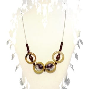 May include: A necklace featuring a thin silver-toned chain, dark brown beads, and four circular translucent tan rings. The two central rings contain white circular inserts decorated with dark brown paisley-like patterns and swirls.