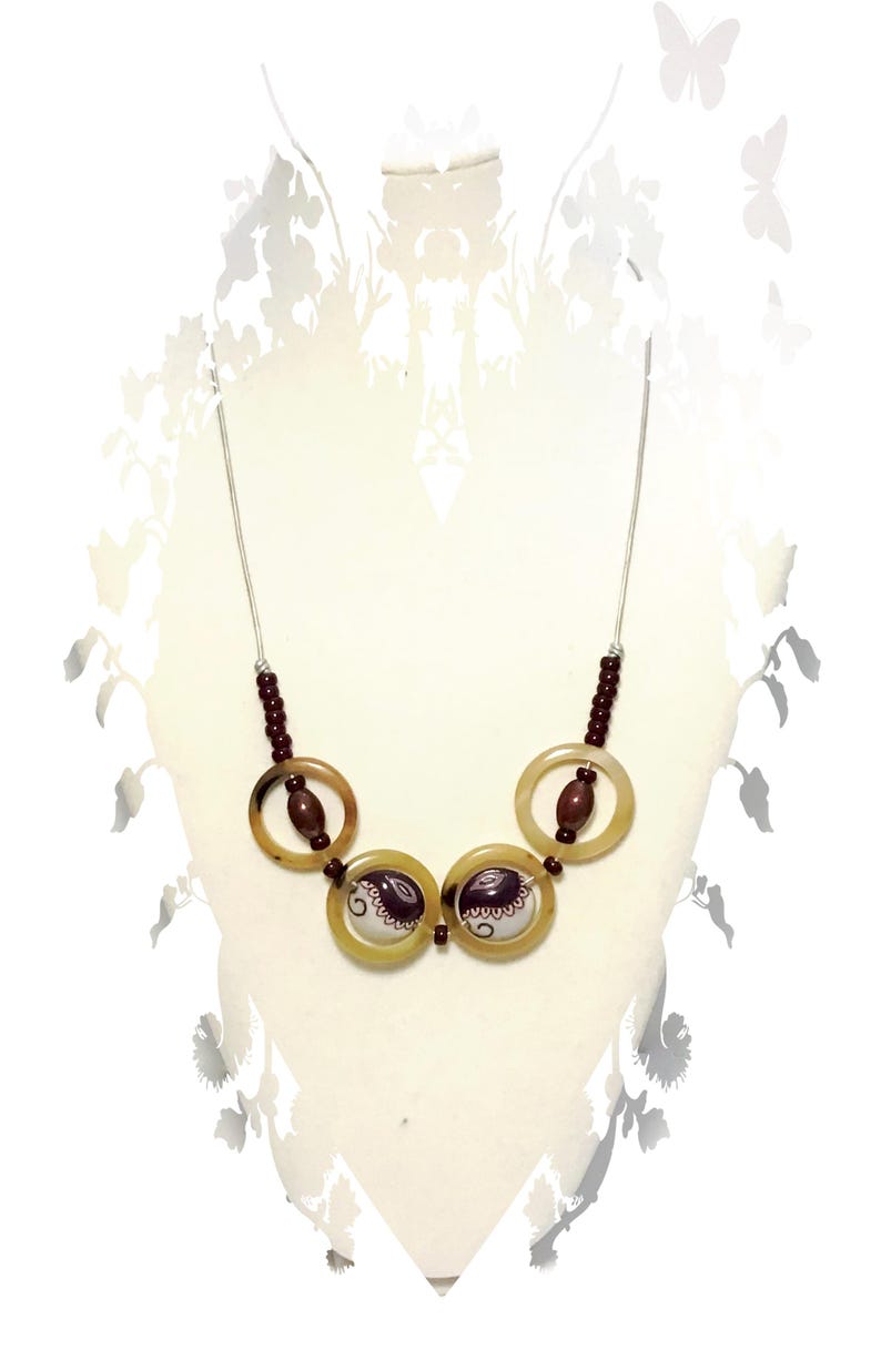 May include: A necklace featuring a thin silver-toned chain, dark brown beads, and four circular translucent tan rings. The two central rings contain white circular inserts decorated with dark brown paisley-like patterns and swirls.