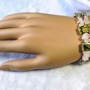 May include: A bracelet featuring rectangular green and brown spotted stones, interspersed with pink, irregular-shaped stones. The bracelet is secured with a silver-colored clasp. The jewelry is displayed on a light-toned surface.