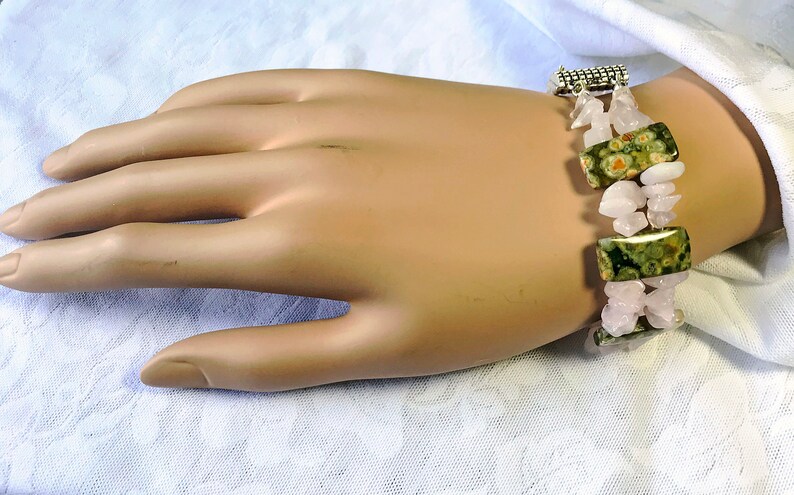 May include: A bracelet featuring rectangular green and brown spotted stones, interspersed with pink, irregular-shaped stones. The bracelet is secured with a silver-colored clasp. The jewelry is displayed on a light-toned surface.