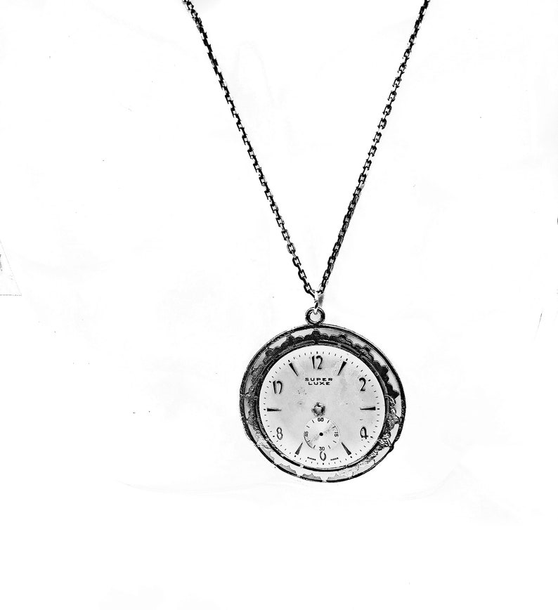 May include: A silver-toned pocket watch pendant with a chain. The watch face is white with black numerals and hands, and the text "SUPER LUXE" is visible. The watch has a small seconds dial and a decorative bezel.