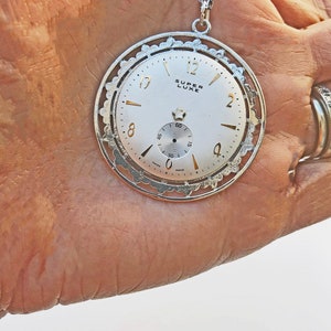 May include: Silver-toned pendant necklace featuring a watch face with the words "SUPER LUXE". The watch face has gold-colored hands and markers, and a small seconds dial. The pendant is held by a silver chain.