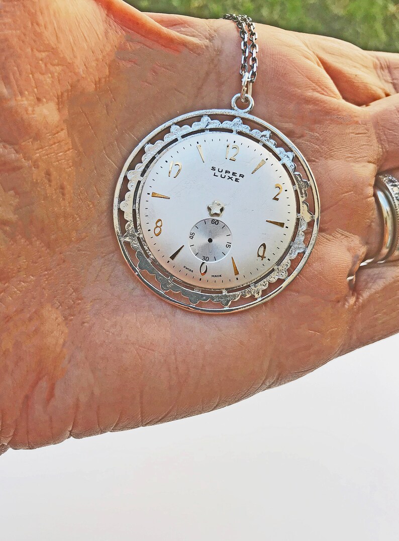 May include: Silver-toned pendant necklace featuring a watch face with the words "SUPER LUXE". The watch face has gold-colored hands and markers, and a small seconds dial. The pendant is held by a silver chain.