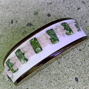May include: A wide bracelet with a white center, framed by a dark brown border. The bracelet features rectangular green stones with a speckled pattern, interspersed with small, light pink stones. A silver-colored clasp secures the bracelet.