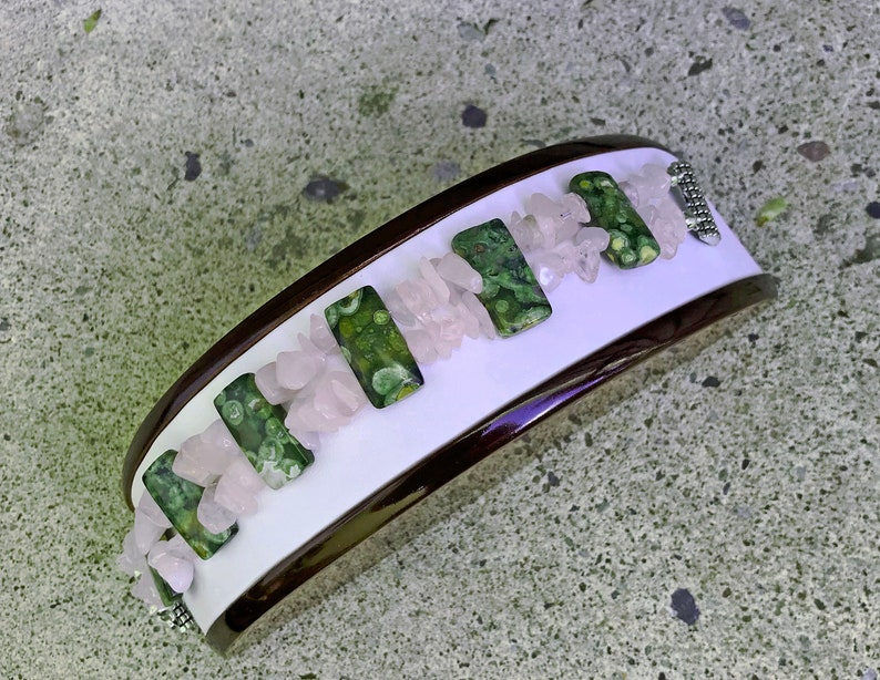 May include: A wide bracelet with a white center, framed by a dark brown border. The bracelet features rectangular green stones with a speckled pattern, interspersed with small, light pink stones. A silver-colored clasp secures the bracelet.