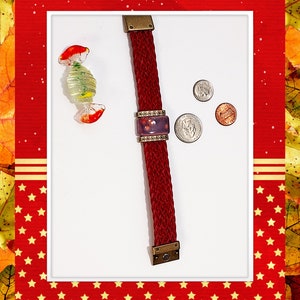 May include: A braided burgundy bracelet with a rectangular purple centerpiece, accented with bronze-colored metal. A glass candy and three coins are also present. The bracelet is approximately 7 inches long.