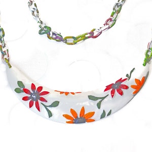 May include: A white necklace with a crescent-shaped pendant adorned with red and orange floral designs and green leaves. The chain is a colorful mix of green, blue, and yellow links.