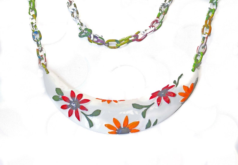 May include: A white necklace with a crescent-shaped pendant adorned with red and orange floral designs and green leaves. The chain is a colorful mix of green, blue, and yellow links.