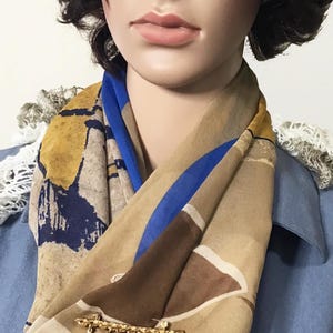 May include: A tan, blue, and brown scarf with a gold brooch. The brooch has coin and pearl charms. The scarf is draped over a mannequin wearing a blue jacket. The text "Shrishti" is visible.