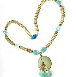May include: A handmade necklace featuring a pendant with a fan-like design and dangling beads. The necklace has light green and blue accents, with a mix of round and faceted beads. The clasp is a blue cylindrical bead.