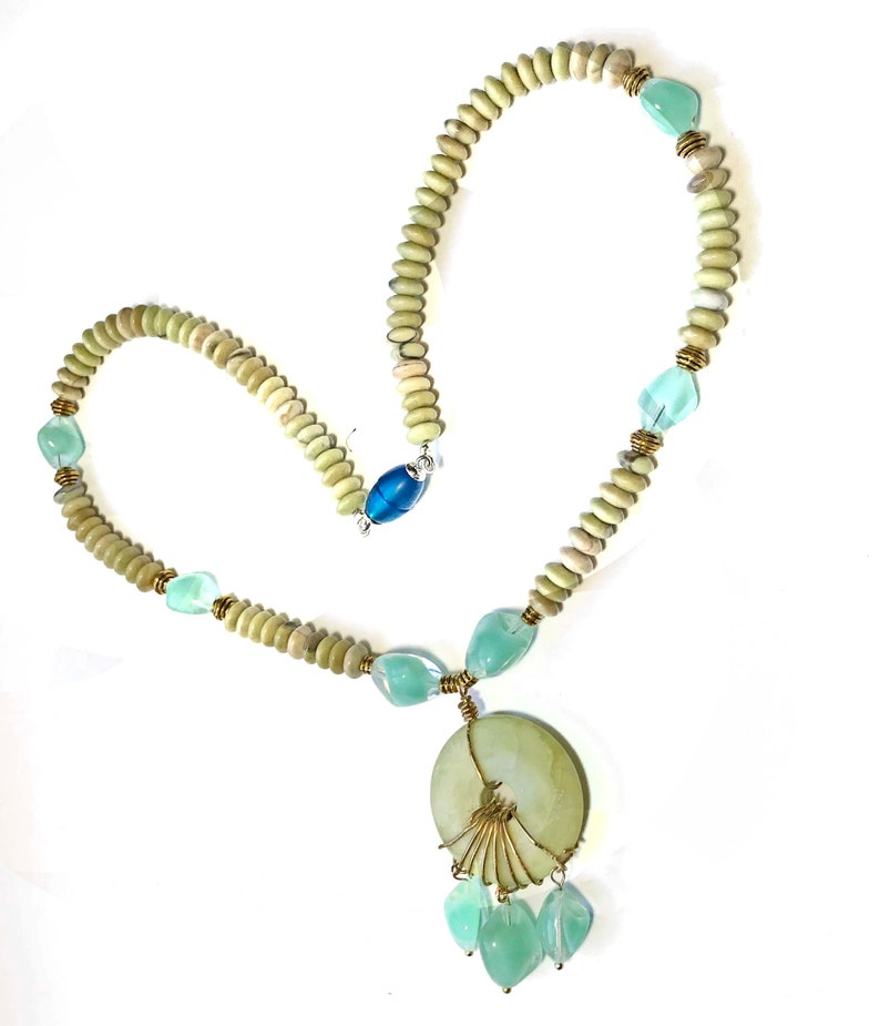 May include: A handmade necklace featuring a pendant with a fan-like design and dangling beads. The necklace has light green and blue accents, with a mix of round and faceted beads. The clasp is a blue cylindrical bead.