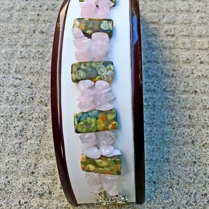 May include: A gemstone bracelet featuring alternating sections of pink rose quartz chips and rectangular green and brown jasper stones. The bracelet is displayed on a white surface with a dark brown border, and has a silver clasp.