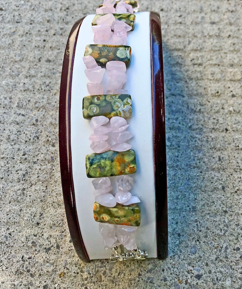 May include: A gemstone bracelet featuring alternating sections of pink rose quartz chips and rectangular green and brown jasper stones. The bracelet is displayed on a white surface with a dark brown border, and has a silver clasp.