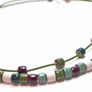 May include: A double-strand necklace featuring cylindrical beads in shades of green, purple, white, and burgundy. The beads are strung on a thin, green cord. The necklace is displayed against a white background.