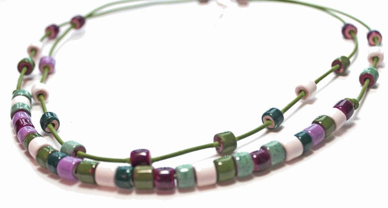 May include: A double-strand necklace featuring cylindrical beads in shades of green, purple, white, and burgundy. The beads are strung on a thin, green cord. The necklace is displayed against a white background.