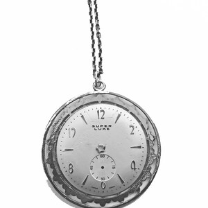 May include: A silver-toned pocket watch with a chain, featuring a white face with black numerals and hands. The watch face has the words "SUPER LUXE" and a smaller seconds dial. The watch has an ornate, textured bezel and is suspended against a white background.