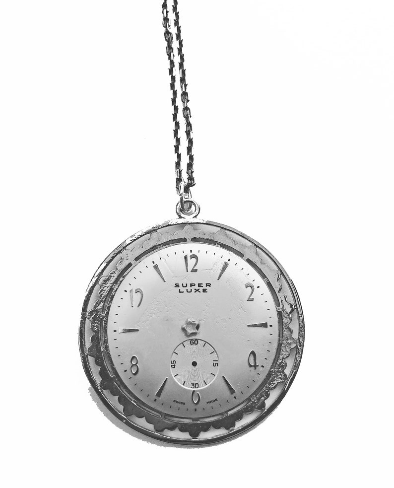 May include: A silver-toned pocket watch with a chain, featuring a white face with black numerals and hands. The watch face has the words "SUPER LUXE" and a smaller seconds dial. The watch has an ornate, textured bezel and is suspended against a white background.