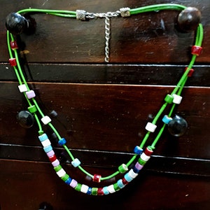 May include: A handmade necklace featuring a vibrant mix of colorful square beads and dark wooden accents. The necklace has a bright green cord and a silver-toned chain. The beads are in shades of red, white, blue, green, and purple.