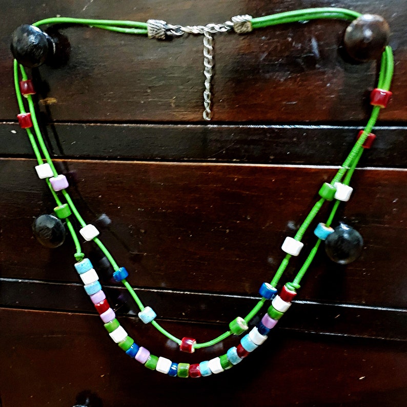 May include: A handmade necklace featuring a vibrant mix of colorful square beads and dark wooden accents. The necklace has a bright green cord and a silver-toned chain. The beads are in shades of red, white, blue, green, and purple.