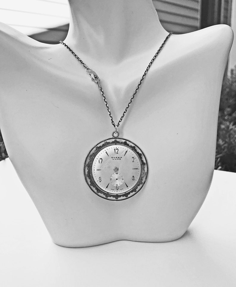 May include: A silver-toned pendant necklace featuring a repurposed vintage watch face. The watch face displays the time and the words "SUPER LUXE". The necklace is displayed on a white mannequin bust.