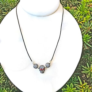 Op de afbeelding: Zwarte koordketting met een schedelhangertje en twee ronde, met kleine, sprankelende stenen versierde accenten. De schedel heeft rode ogen. De ketting wordt getoond op een witte buste tegen een achtergrond van groen gebladerte.