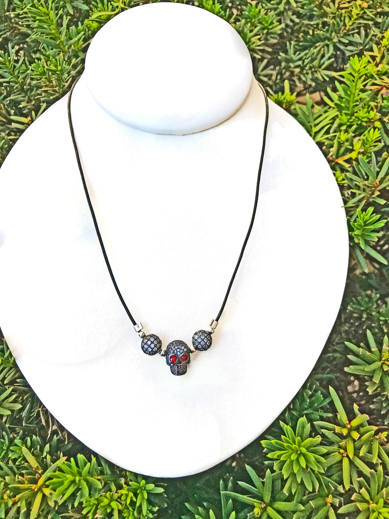 Op de afbeelding: Zwarte koordketting met een schedelhangertje en twee ronde, met kleine, sprankelende stenen versierde accenten. De schedel heeft rode ogen. De ketting wordt getoond op een witte buste tegen een achtergrond van groen gebladerte.