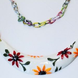 May include: A white, crescent-shaped necklace pendant adorned with red and orange floral designs and green leaves. The pendant is connected to a colorful chain necklace. The chain links are painted with a variety of colors.
