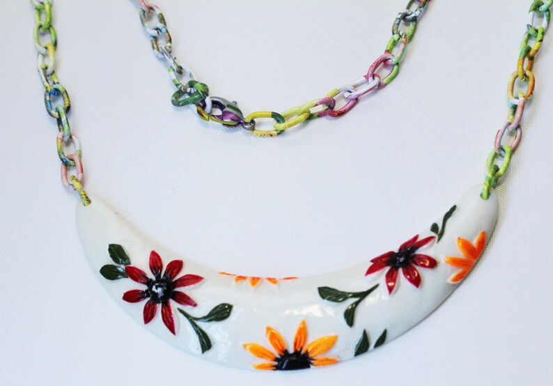 May include: A white, crescent-shaped necklace pendant adorned with red and orange floral designs and green leaves. The pendant is connected to a colorful chain necklace. The chain links are painted with a variety of colors.