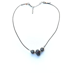 Op de afbeelding: Zwarte koordketting met een schedelhangertje met rode accenten en twee met kristal bezette kralen. De ketting heeft een zilverkleurige ketting en sluiting, tegen een witte achtergrond.