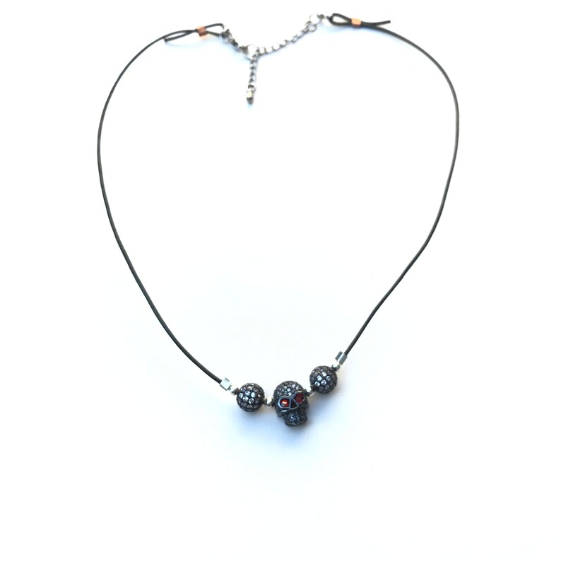 Op de afbeelding: Zwarte koordketting met een schedelhangertje met rode accenten en twee met kristal bezette kralen. De ketting heeft een zilverkleurige ketting en sluiting, tegen een witte achtergrond.