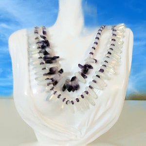 May include: A white necklace displayed on a white bust. The necklace features clear, elongated beads and dark purple, irregularly shaped stones. The necklace is layered, with a strand of small purple beads. The background is a blurred blue sky.