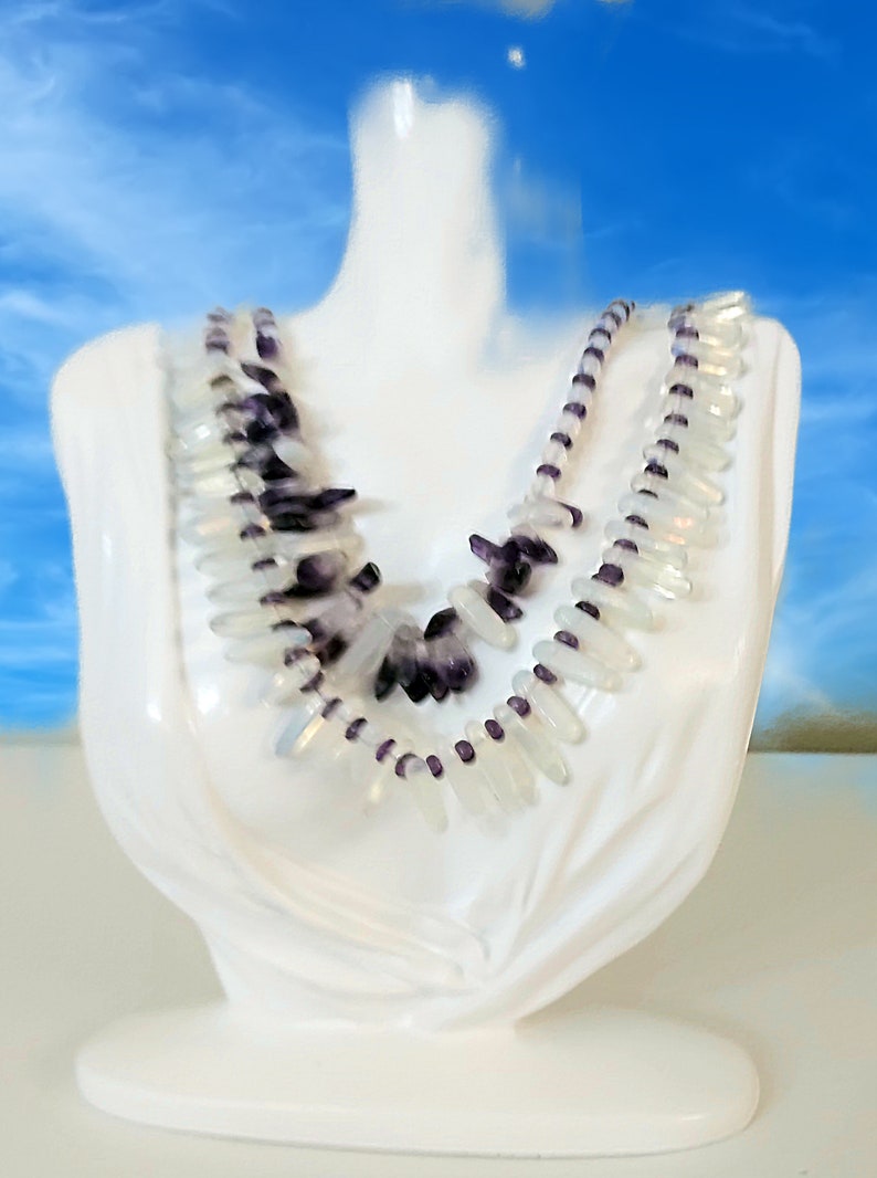 May include: A white necklace displayed on a white bust. The necklace features clear, elongated beads and dark purple, irregularly shaped stones. The necklace is layered, with a strand of small purple beads. The background is a blurred blue sky.