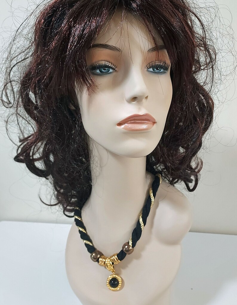 May include: A black and gold necklace with a pendant featuring a black stone. The necklace has a braided design with gold accents and is displayed on a mannequin. The pendant is round and has a gold frame.