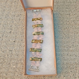 May include: A handmade bracelet featuring alternating sections of pink rose quartz chips and rectangular, patterned stones in shades of green, brown, and gold. The bracelet is secured with a silver-toned clasp and is presented in a small, open cardboard box.