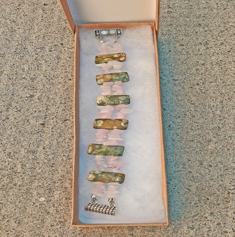 May include: A handmade bracelet featuring alternating sections of pink rose quartz chips and rectangular, patterned stones in shades of green, brown, and gold. The bracelet is secured with a silver-toned clasp and is presented in a small, open cardboard box.