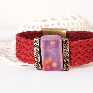 May include: Red braided leather bracelet with a rectangular purple and orange ceramic centerpiece. The centerpiece is framed by bronze-colored metal with a swirl design. The bracelet has a bronze-colored magnetic clasp.