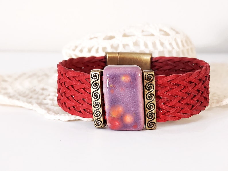 May include: Red braided leather bracelet with a rectangular purple and orange ceramic centerpiece. The centerpiece is framed by bronze-colored metal with a swirl design. The bracelet has a bronze-colored magnetic clasp.