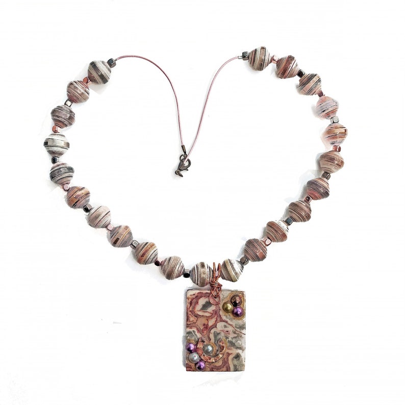 May include: A handmade necklace featuring paper beads in shades of brown, gray, and white. The necklace is strung on a pink cord, forming a heart shape. A rectangular pendant with a patterned design and decorative accents hangs from the necklace.
