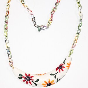 May include: A colorful necklace featuring a white, curved pendant adorned with red and orange floral designs and green leaves. The necklace has a chain with links in various colors, and a clasp.
