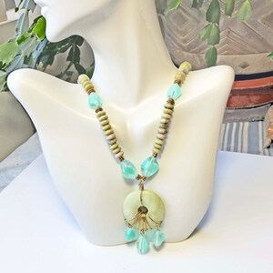 May include: A handcrafted necklace featuring a large, pale green circular pendant with a gold wire design and dangling aqua-colored beads. The necklace has light green and tan wooden beads and aqua-colored glass beads.
